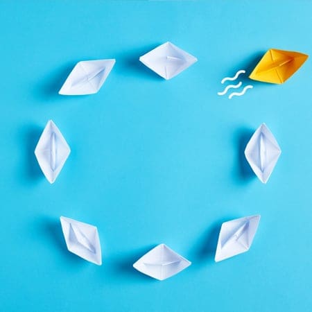 A group of seven white paper boats arranged in a circular formation on a blue background. An eighth paper boat, colored yellow, is outside the circle with three wavy lines near it, indicating movement away from the group. A group of seven white paper boats arranged in a circular formation on a blue background. An eighth paper boat, colored yellow, is outside the circle with three wavy lines near it, indicating movement away from the group.
