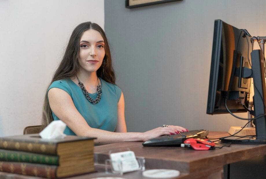 A woman with long hair sits at a desk, typing on a keyboard connected to a desktop computer. She wears a teal top and a beaded necklace. The desk has books, tissues, and some office supplies on it. The background features a gray and white wall.