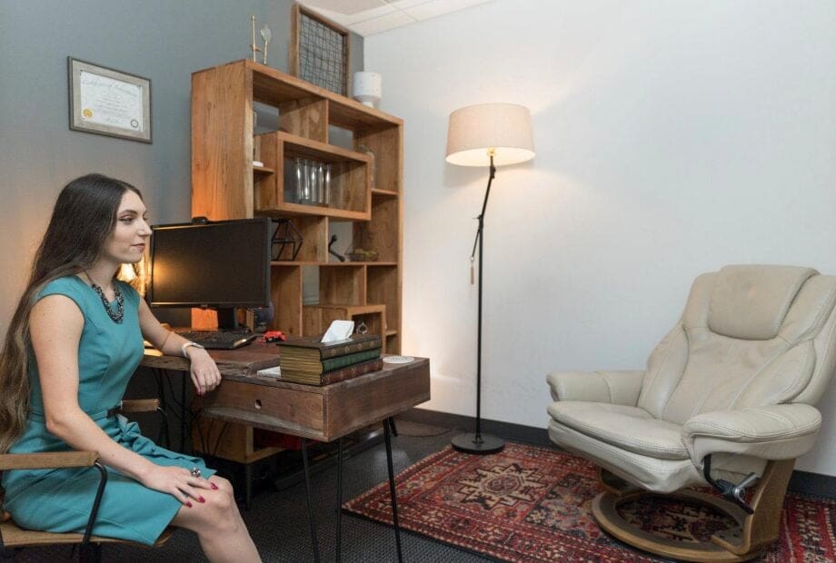 A woman with long hair, dressed in a teal dress, sits at a wooden desk in an office setup. The desk holds a computer monitor, books, and a box of tissues. Adjacent to her is an empty beige recliner, a floor lamp, and a wooden shelf filled with various items.