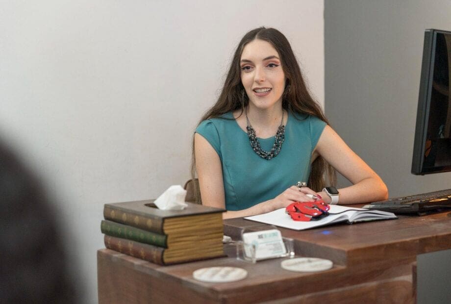 A woman with long hair, wearing a teal dress and a necklace, is sitting at a wooden desk. The desk has books, tissues, a computer monitor, and small items. She is smiling, with an open notebook and a pen in front of her.
