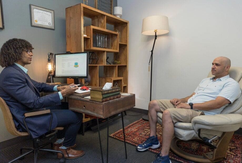 A man sits in a comfortable chair during a counseling session with a therapist in an office. The therapist is seated at a wooden desk with a computer screen displaying charts. Books and a tissue box are on the desk, and a framed certificate hangs on the wall.
