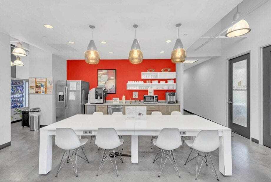 A modern break room with a long white table and white chairs. The back wall is painted orange with shelves holding kitchen items. There are stainless steel appliances, including a refrigerator and coffee machines. Pendant lights hang above the table.