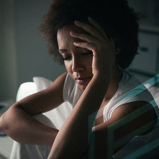 A person sits with their eyes closed, resting their head on their hand, and appears to be deep in thought or feeling emotional. They are in a dimly lit room, wearing a white sleeveless top.