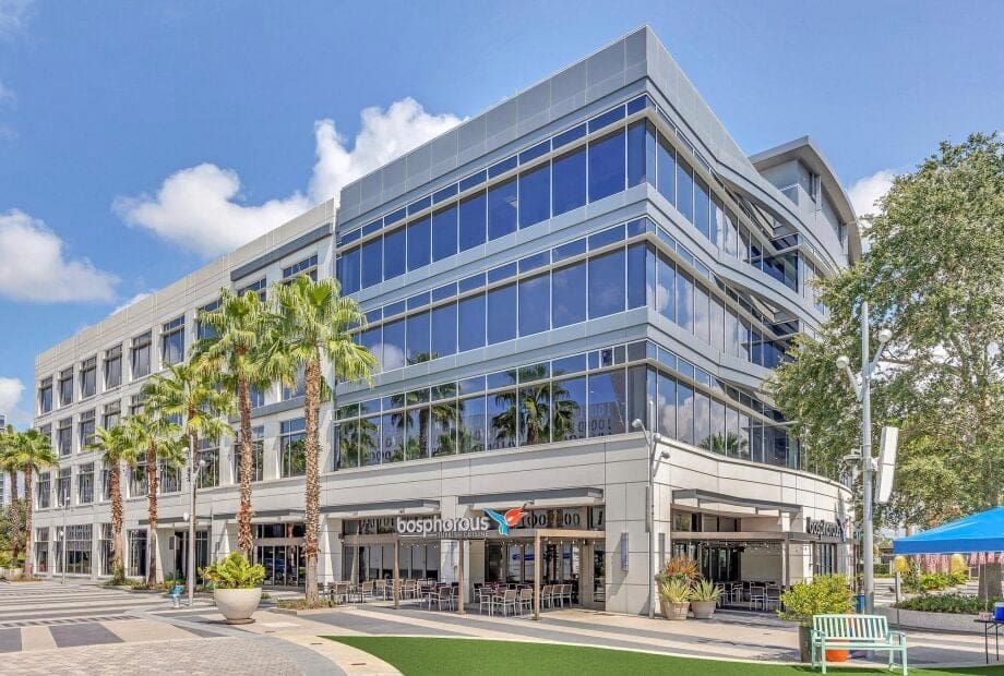 A modern, four-story office building with large glass windows and geometric architectural features. The ground floor hosts businesses, including a restaurant called Bosphorus with outdoor seating. Tall palm trees and a neatly maintained plaza surround the building.