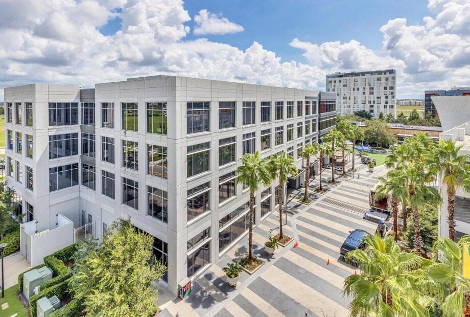 A modern, multi-story office building with large windows is surrounded by palm trees and landscaped walkways. A striped pedestrian area and a parking lot with cars are visible, along with another building in the background under a partly cloudy sky.