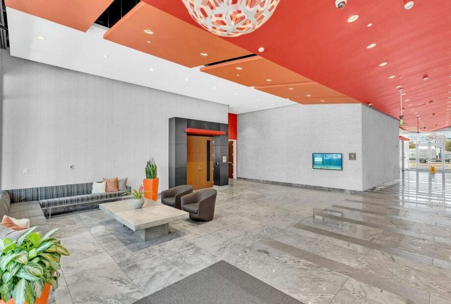 A modern lobby with a sleek design. It features a marble floor, an elevator with a wooden door, and seating that includes a gray sectional sofa, two brown chairs, and a low coffee table. Orange ceiling panels and hanging lights add color to the space.