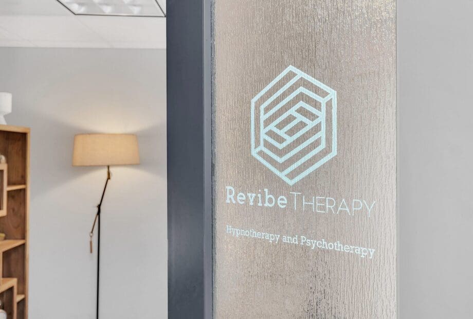 A frosted glass door with the text "Revibe Therapy" and "Hypnotherapy and Psychotherapy" is featured in an office. In the background, there is a wooden shelf and a lit floor lamp with a beige shade. The office has a calm and welcoming atmosphere.