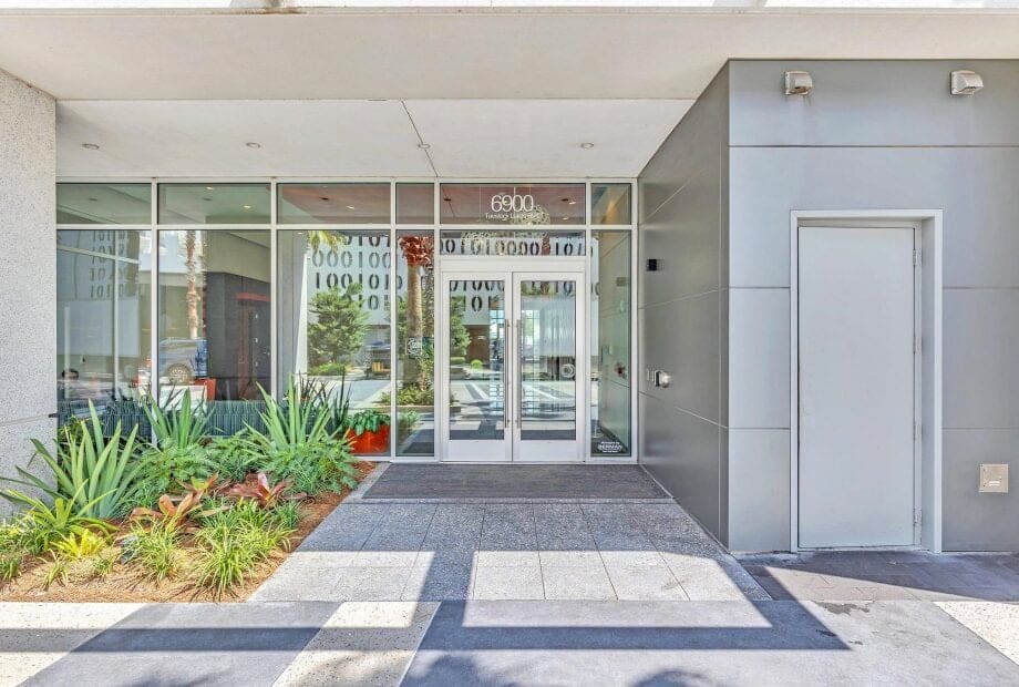 Front entrance of a modern building with the address number "6800" displayed above the glass double doors. The exterior features a mix of concrete and metal surfaces, with a manicured garden area to the left of the entrance and a closed utility door to the right.