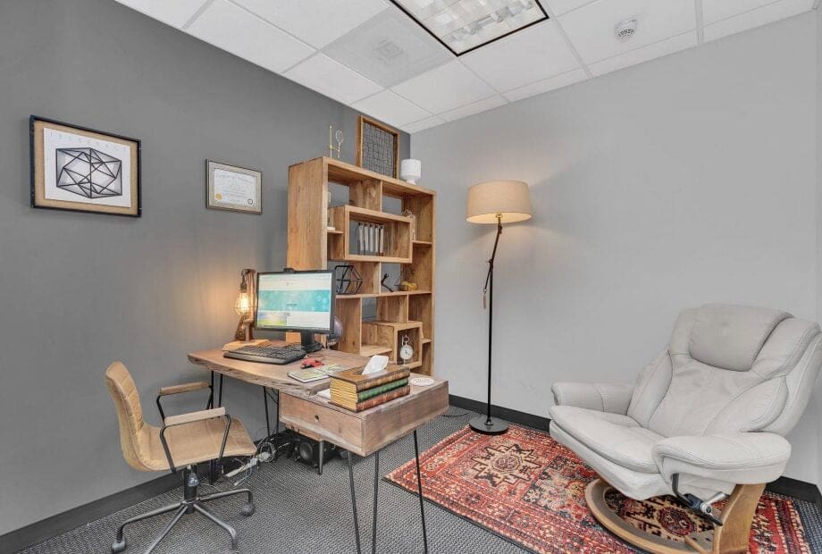 A cozy office space with a wooden desk holding a computer, lamp, and various books. A cushioned chair sits nearby on a patterned rug. Shelves on the wall contain more books and decor. A framed certificate and artwork decorate the gray walls under a ceiling light.