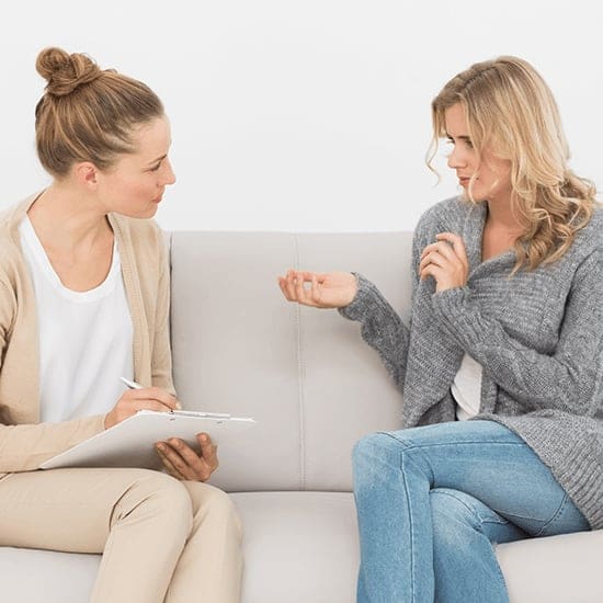Two women are sitting on a couch facing each other. One woman, holding a clipboard and pen, appears to be a therapist or counselor, while the other woman, who is gesturing with her hands, seems to be speaking or explaining something. Both look engaged in conversation. Two women are sitting on a couch facing each other. One woman, holding a clipboard and pen, appears to be a therapist or counselor, while the other woman, who is gesturing with her hands, seems to be speaking or explaining something. Both look engaged in conversation.