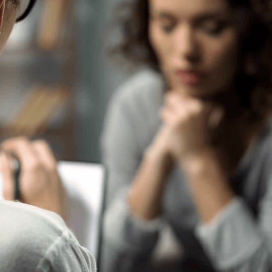 A person with blurry features, wearing a gray sweater, is sitting with their hands clasped near their face, appearing to be in deep thought or prayer. Another person, out of focus, sits across from them, holding a notebook and pen. A person with blurry features, wearing a gray sweater, is sitting with their hands clasped near their face, appearing to be in deep thought or prayer. Another person, out of focus, sits across from them, holding a notebook and pen.