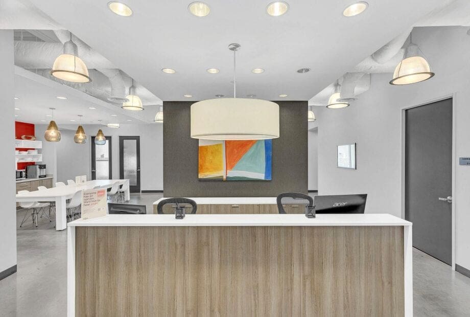 A modern, open office space with a front reception desk made of light wood and a large pendant light above it. Two black chairs sit behind the desk. In the background, there's colorful abstract wall art and a dining area with tables and chairs.