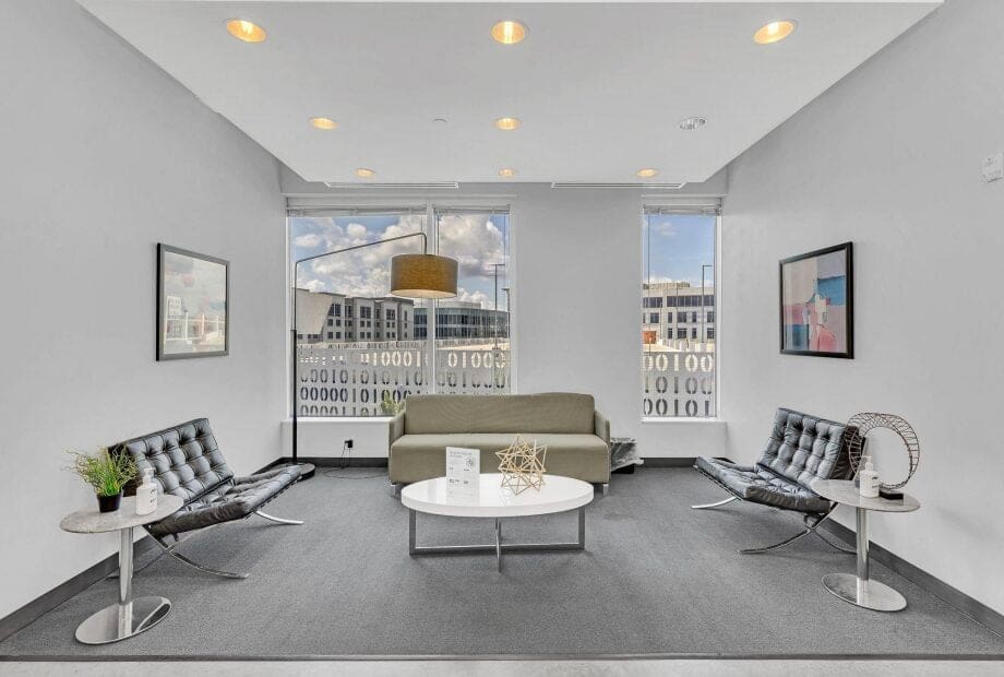 A modern waiting room with two black leather chairs, a gray sofa, and a white coffee table. There are two abstract paintings on the walls, indoor plants, and large windows with partial city views. The room is lit by recessed ceiling lights.