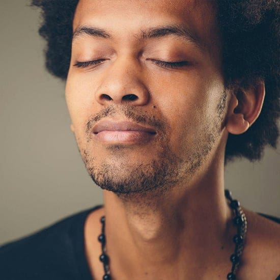 A person with closed eyes, slight smile, and relaxed expression stands against a neutral background. They have short curly hair, a mustache, and a goatee, and are wearing a beaded necklace and a dark shirt. A person with closed eyes, slight smile, and relaxed expression stands against a neutral background. They have short curly hair, a mustache, and a goatee, and are wearing a beaded necklace and a dark shirt.