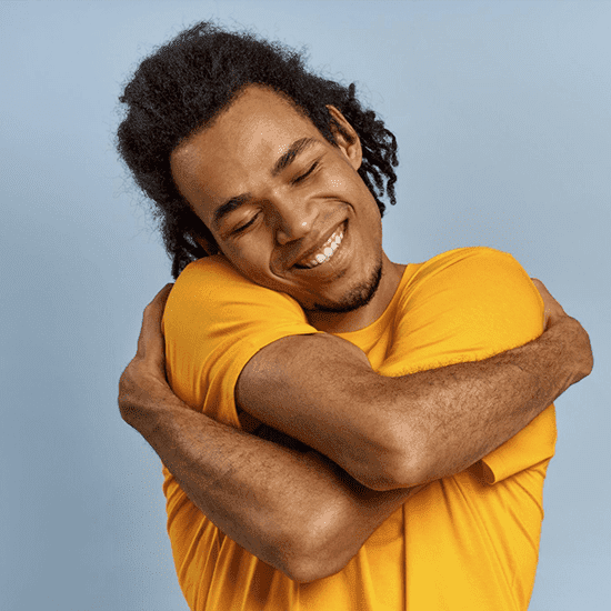 A person with medium-length curly hair wearing an orange shirt is smiling with their eyes closed while hugging themselves. They stand against a plain light blue background. A person with medium-length curly hair wearing an orange shirt is smiling with their eyes closed while hugging themselves. They stand against a plain light blue background.