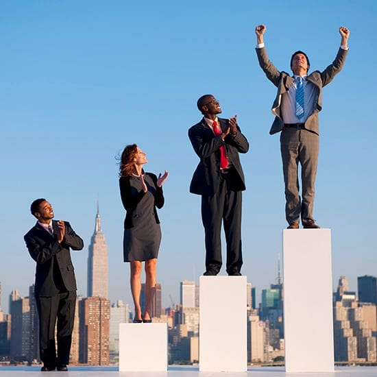 Four business professionals stand on different height podiums against a city skyline. The person on the tallest podium raises their arms in victory, the others applaud. Two men wear suits, one man and one woman wear business attire. Four business professionals stand on different height podiums against a city skyline. The person on the tallest podium raises their arms in victory, the others applaud. Two men wear suits, one man and one woman wear business attire.