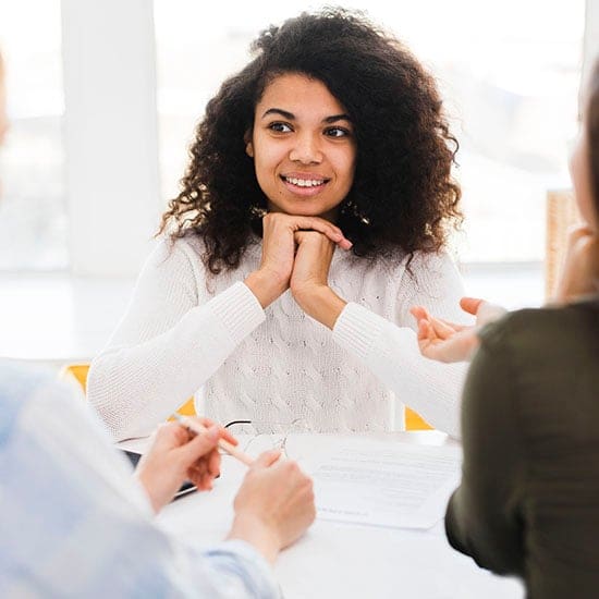 A woman with curly hair wearing a white sweater is sitting at a table with her hands clasped under her chin, smiling, and listening intently to two other people who are partially visible and sitting across from her. Documents on the table suggest topics of life coaching may be under discussion. A woman with curly hair wearing a white sweater is sitting at a table with her hands clasped under her chin, smiling, and listening intently to two other people who are partially visible and sitting across from her. Documents on the table suggest topics of life coaching may be under discussion.