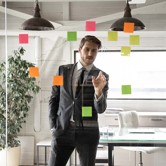 A man in a suit stands behind a glass wall covered with colorful sticky notes, pointing at one of the notes. He is in a modern office setting with large windows, a potted plant, and a desk visible in the background. A man in a suit stands behind a glass wall covered with colorful sticky notes, pointing at one of the notes. He is in a modern office setting with large windows, a potted plant, and a desk visible in the background.