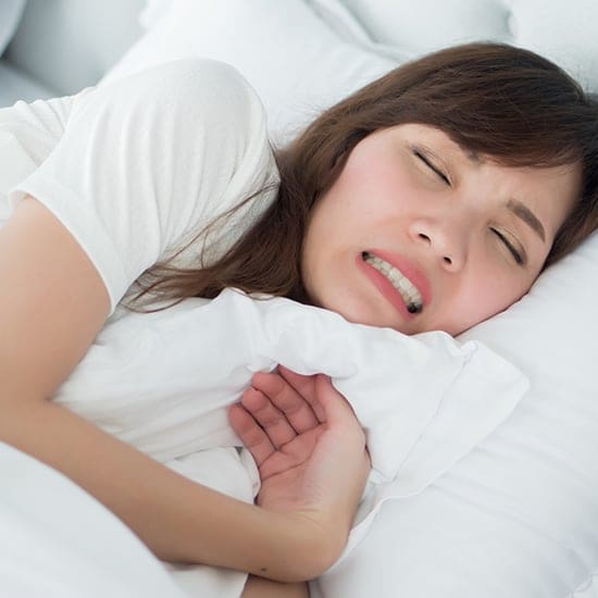 A person with long brown hair lies in bed on a white pillow, appearing to be in discomfort or pain, with eyes closed and teeth clenched. One hand is tucked under the pillow while the other clutches part of the white blanket, a scene indicative of someone struggling with mental disorders. They wear a white shirt. A person with long brown hair lies in bed on a white pillow, appearing to be in discomfort or pain, with eyes closed and teeth clenched. One hand is tucked under the pillow while the other clutches part of the white blanket, a scene indicative of someone struggling with mental disorders. They wear a white shirt.