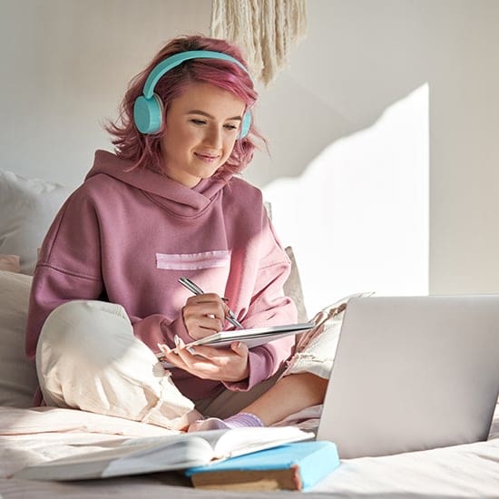 A person with pink hair is sitting on a bed, wearing teal headphones and a pink hoodie. They are taking notes in a notebook while looking at an open laptop. Sunlight streams through a window, and a couple of books lie nearby. A person with pink hair is sitting on a bed, wearing teal headphones and a pink hoodie. They are taking notes in a notebook while looking at an open laptop. Sunlight streams through a window, and a couple of books lie nearby.