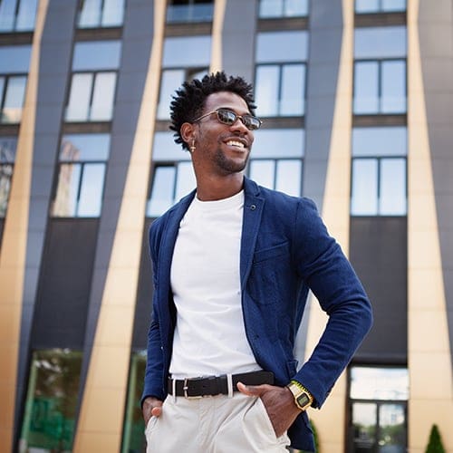 A man with sunglasses is smiling and standing in front of a modern building with gold and black wavy architecture. He is wearing a white t-shirt, blue blazer, beige pants with a black belt, and a yellow watch. His hands are in his pockets.