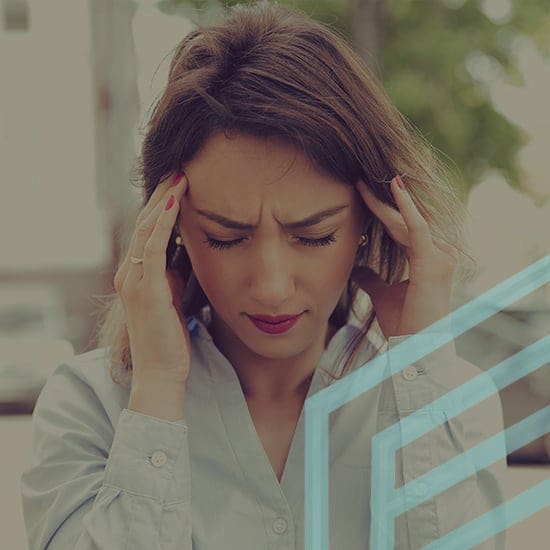 A woman with long hair wears a light blue shirt. She looks distressed with furrowed brows and eyes closed, holding her temples with both hands. The background and surrounding details appear slightly blurred.