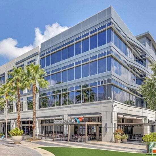 A modern multi-story commercial building with large glass windows. The first floor has a restaurant named "Bosphorous" with outdoor seating. Palm trees and decorative plants surround the entrance. The sky is clear with a few clouds.