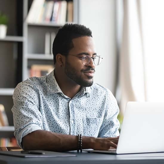 A person wearing glasses and a patterned shirt is sitting at a desk while looking at an open laptop. A bookcase and window are visible in the background.