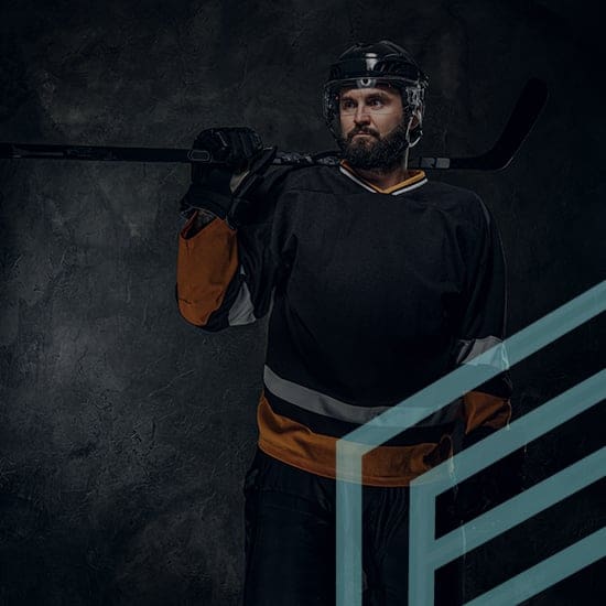 Hockey player standing against a dark background, holding a hockey stick over his shoulder. He is wearing a black jersey with orange accents, protective gear, and a helmet. His expression is serious and focused.