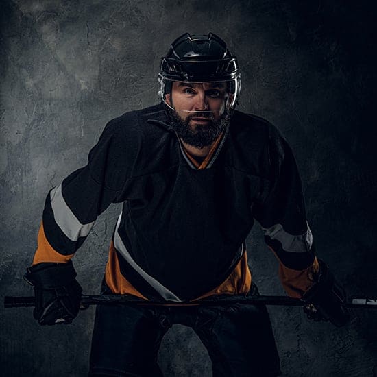 A hockey player wearing a black and orange uniform and a helmet, holding a hockey stick, poses against a dark, textured background. The player has a focused expression and a prominent beard.