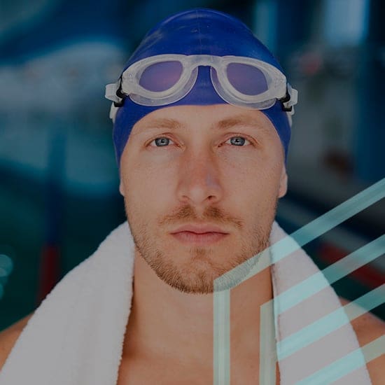 A swimmer wearing a blue swim cap and goggles rests with a white towel around his shoulders. He is looking directly at the camera with a focused expression. The blurred background suggests an indoor pool environment.