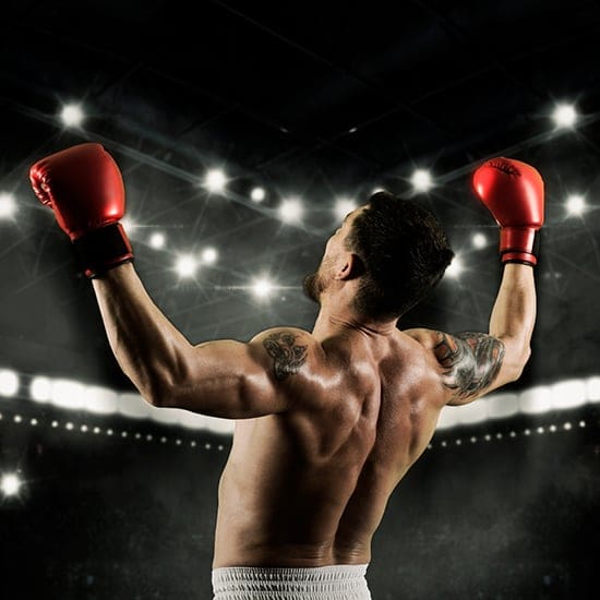 A male boxer wearing red gloves and white shorts stands with his back to the camera, arms raised in victory under bright stadium lights. The background is dark with illuminated spotlights and a blurred crowd, emphasizing the triumph and excitement.