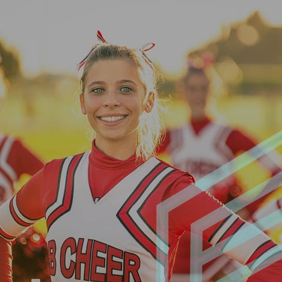 A cheerleader in a red and white uniform, standing outdoors and smiling at the camera. Two other cheerleaders are slightly blurred in the background. The image is bright and appears to be taken during sunset.