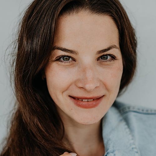 Close-up of a woman with long brown hair and light skin, smiling gently at the camera. She has minimal makeup and is wearing a light blue denim jacket. The background is simple and out of focus, emphasizing her natural features.