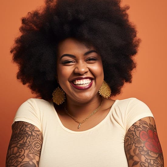 A person with a voluminous afro hairstyle smiles brightly against an orange background. They are wearing a beige top, large intricate gold earrings, a necklace, and have tattoos covering their arms.