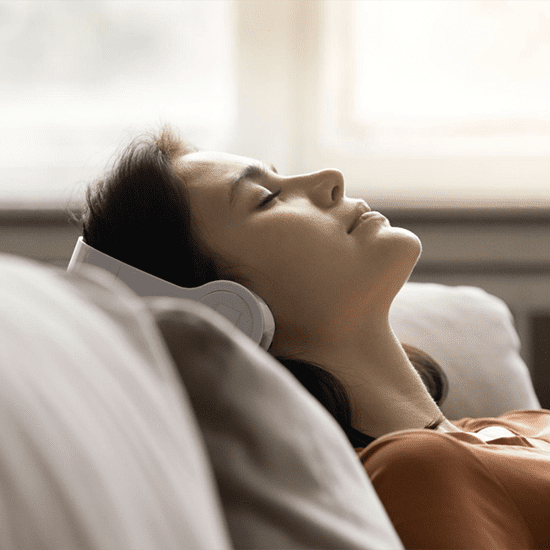 A woman with dark hair lies on a couch with her eyes closed, wearing white over-ear headphones. She appears relaxed and serene, and soft daylight filters through a window in the background.