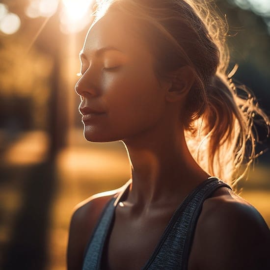 A woman with closed eyes, standing outdoors in soft golden sunlight. She has a serene expression, and her hair is tied back in a loose ponytail. She is wearing a sleeveless top, and the background features blurred trees and warm light.