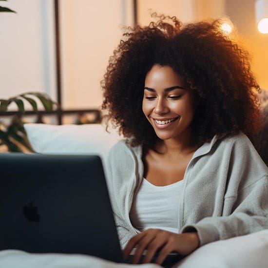 A woman with curly hair is seated on a couch, smiling at the screen of her laptop. She is wearing a cozy, light-colored zip-up jacket over a white top. The background is softly lit, giving the scene a warm, relaxed ambiance.