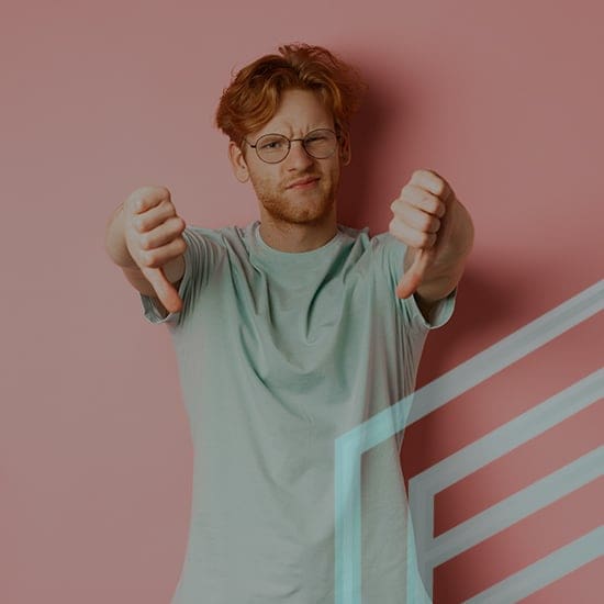 A person with red hair and glasses stands against a pink background, wearing a light grey T-shirt. They are squinting one eye and giving a double thumbs-down gesture.