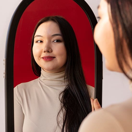 A woman with long dark hair and a beige turtleneck top is smiling and looking into a mirror, reflecting her face. The background of the mirror is red, contrasting with the white wall behind her.