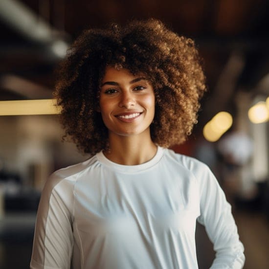 A woman with curly hair smiles warmly at the camera. She is wearing a white athletic shirt and standing in what appears to be a gym or fitness center. The background is softly blurred with some warm lights creating a cozy atmosphere.