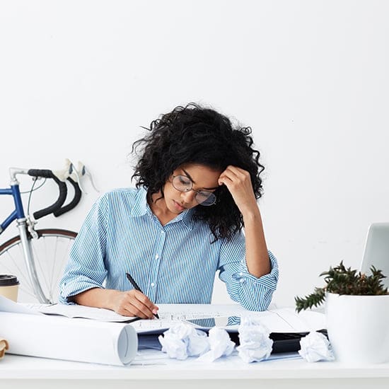 A woman with curly hair and glasses, wearing a blue striped shirt, sits at a desk with a thoughtful expression, writing on a notepad. The desk has crumpled paper, a coffee cup, and architectural blueprints. A bicycle is in the background.