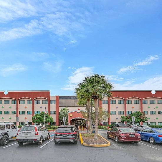 A modern, three-story building with a red and cream exterior is shown, featuring arched windows and an entrance flanked by palm trees. Several cars are parked in the foreground parking lot under a partly cloudy blue sky.