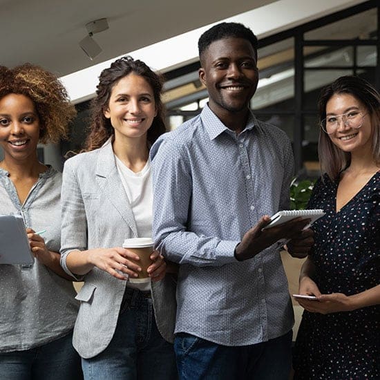 A group of four diverse people standing together, all smiling at the camera. They are holding notepads, coffee cups, and tablets. The background features an office setting with glass walls and modern decor.