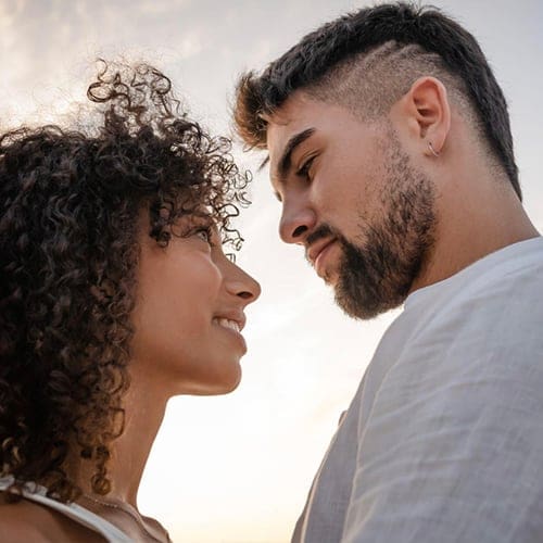 A couple stands closely facing each other against a soft, cloudy sky. The woman on the left has curly hair and smiles, and the man on the right has short, styled hair with an undercut and a beard. They seem to be sharing an intimate moment, looking into each other’s eyes. A couple stands closely facing each other against a soft, cloudy sky. The woman on the left has curly hair and smiles, and the man on the right has short, styled hair with an undercut and a beard. They seem to be sharing an intimate moment, looking into each other's eyes.