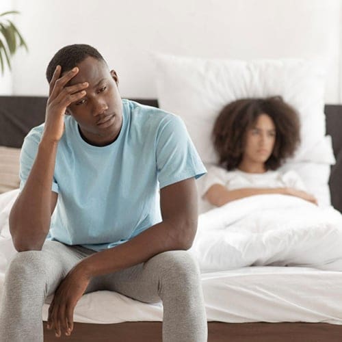 A man in a blue shirt and gray pants sits on the edge of a bed, looking pensive with his hand on his forehead. In the background, a person with curly hair lies in bed, partially covered by white bedding, looking away from the man. The setting appears to be a bedroom. A man in a blue shirt and gray pants sits on the edge of a bed, looking pensive with his hand on his forehead. In the background, a person with curly hair lies in bed, partially covered by white bedding, looking away from the man. The setting appears to be a bedroom.