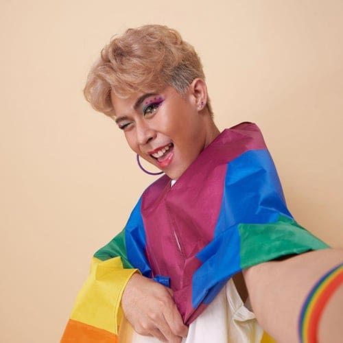 A person with blonde hair and colorful makeup is smiling and winking at the camera. They are draped in a rainbow flag, holding one end of it, and wearing a rainbow wristband. The background is a plain beige wall. A person with blonde hair and colorful makeup is smiling and winking at the camera. They are draped in a rainbow flag, holding one end of it, and wearing a rainbow wristband. The background is a plain beige wall.