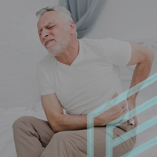 A bearded elderly man in a white t-shirt and beige pants sitting on the edge of a bed, grimacing in pain while clutching his lower back with one hand. The background is a light-colored bedroom with a curtain visible.