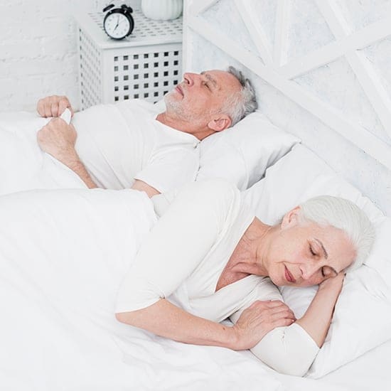 An elderly couple sleeps in a white bed, wearing white clothes. The man is on his back, while the woman lies on her side facing away from him. There is a small nightstand with an alarm clock next to the bed. The room is peaceful and bright.