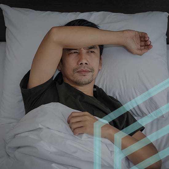 A man wearing a dark shirt lies awake in bed with his left arm resting across his forehead. He appears contemplative or unable to sleep, looking upward. The bed has white sheets and pillows, and a translucent geometric pattern overlays a corner of the image.